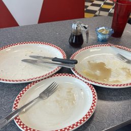 This is an indication how good everything was! - A diner table features three empty plates with red and white checkered borders, along with a fork and knife. A small pitcher of syrup, a packet of butter, and a red drinking glass are also on the table. The background shows red and white booth seating and a checkered floor, indicating a classic American diner environment.