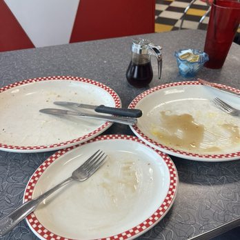 Ze's Diner - This is an indication how good everything was! - A diner table features three empty plates with red and white checkered borders, along with a fork and knife. A small pitcher of syrup, a packet of butter, and a red drinking glass are also on the table. The background shows red and white booth seating and a checkered floor, indicating a classic American diner environment. - Diners Near Me - Eagan, Minnesota