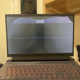 Before fixing - A Dell laptop with a visibly cracked screen, showing a black display with damage at the top left corner and some distortion. The keyboard is illuminated with orange backlighting, and the laptop is placed on a surface in a room with a wooden shelf and a door in the background. The screen damage affects the display quality and visibility.