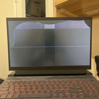 EzFix - Before fixing - A Dell laptop with a visibly cracked screen, showing a black display with damage at the top left corner and some distortion. The keyboard is illuminated with orange backlighting, and the laptop is placed on a surface in a room with a wooden shelf and a door in the background. The screen damage affects the display quality and visibility. - Mobile Phone Repair Near Me - Glendora, California