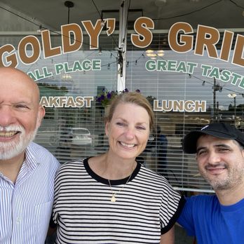 Goldy's Grill - Three people are taking a selfie in front of the window of Goldy's Grill, a restaurant with a sign displaying 'All Place, Great Taste, Breakfast, Lunch.' The group includes an older man with a white beard and striped shirt, a woman with short hair and a striped top, and a man wearing a blue shirt and black cap. The background shows the restaurant's interior through the window, with reflections of parked cars and outdoor lighting. The image captures a casual, friendly moment during their visit. - American Near Me - Park Ridge, Illinois