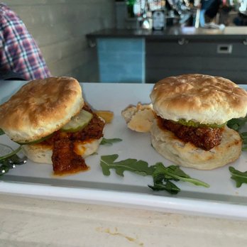 The Local - Two small sandwiches with pickles and sauce are placed on a white rectangular plate, garnished with greens. The sandwiches appear to have soft buns and contain a filling with sauce and pickles. The background shows a bar or restaurant interior with a person wearing a plaid shirt partially visible on the left and bar equipment in the distance. - Cocktail Bars Near Me - Babylon, New York