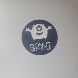 A logo displays a cartoon monster with one eye, horns, and arms, inside a dark circle. The monster has a round body, a single eye, and a wide mouth with small teeth. Below the monster, the text reads "Donut Monster" in a playful, uppercase font. The logo is printed on a white background, likely on a box or packaging for a donut shop.