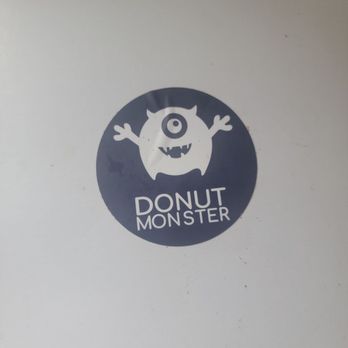 Donut Monster Third Ward - A logo displays a cartoon monster with one eye, horns, and arms, inside a dark circle. The monster has a round body, a single eye, and a wide mouth with small teeth. Below the monster, the text reads "Donut Monster" in a playful, uppercase font. The logo is printed on a white background, likely on a box or packaging for a donut shop. - Donuts Near Me - Milwaukee, Wisconsin