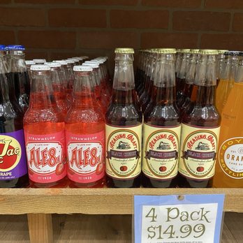 Rocket Fizz - Insane soda pop prices - The image shows a selection of bottled soft drinks on a wooden shelf, including Ale8 Strawberry and Black Cherry soda. The bottles are arranged in rows, with Ale8 bottles on the left and Black Cherry bottles on the right. A sign below indicates the price for a 4-pack of these beverages is $14.99. The background features a brick wall, and the bottles are sealed with metal caps. - Candy Stores Near Me - Bloomington, Minnesota