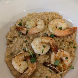 A dish of shrimp risotto is presented on a white, oval-shaped plate. The risotto appears creamy with visible grains of rice and small green herbs. Four cooked shrimp are arranged on top, garnished with microgreens. The background shows a marble countertop, indicating a restaurant or home setting.