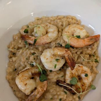 Mattarello - A dish of shrimp risotto is presented on a white, oval-shaped plate. The risotto appears creamy with visible grains of rice and small green herbs. Four cooked shrimp are arranged on top, garnished with microgreens. The background shows a marble countertop, indicating a restaurant or home setting. - Italian Near Me - Markham, Ontario