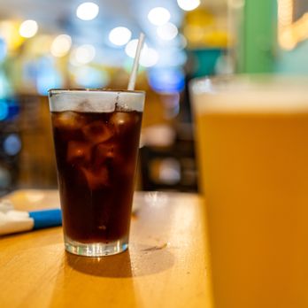 Koco's Pub - Coke Zero® ($3) - A tall, clear glass filled with Coke Zero® containing ice cubes and a straw is positioned on a wooden table. The background features a blurred, colorful restaurant interior with bright lights and various colors, creating a lively atmosphere. The focus is on the beverage, with the surrounding environment out of focus. - Pubs Near Me - Baltimore, Maryland