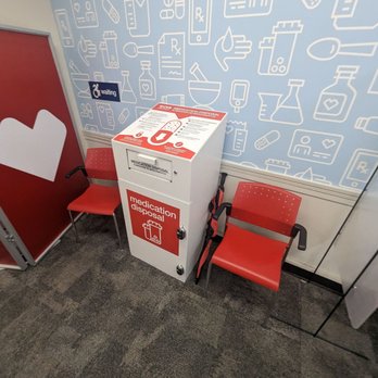 CVS Pharmacy - A medication disposal kiosk is placed in a healthcare or pharmacy environment, flanked by two red chairs with armrests. The kiosk has instructions on top and a slot for disposing of medications. The background features a blue wall decorated with white medical-themed icons, including bottles, pills, and medical devices, indicating a medical or pharmacy setting. - Drugstores Near Me - Watertown, Massachusetts