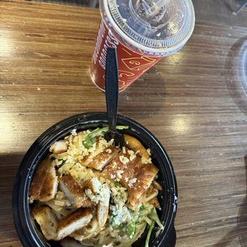 Noodles and Company - A black bowl contains Asian-style noodle salad with grilled chicken pieces, shredded cheese, and vegetables, accompanied by a soft drink in a plastic cup with a straw. - Noodles Near Me - Tempe, Arizona
