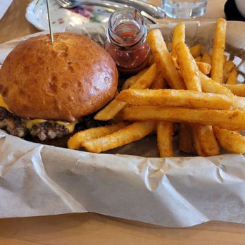 Leader - Burger and fries with ketchup. - A cheeseburger with a toasted bun, melted cheese, and a beef patty is placed on a tray lined with parchment paper. Next to it, there is a portion of French fries with a small jar of ketchup. The tray is on a wooden table, with utensils, a glass of water, and a plate visible in the background. - Scandinavian Near Me - Cambridge, Minnesota