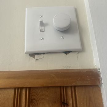 Starr Electrical Services - Two holes in my wall made by Starr electric - A white electrical outlet with a switch and a cover plate is installed on a wall above a wooden door frame. The wall shows damage and peeling paint beneath the outlet, exposing the underlying material. The outlet appears to be a standard electrical component, with the switch in the 'on' position. The surrounding area includes a section of painted wall and a wooden door frame at the bottom of the image. - Electricians Near Me - Hermon, Maine