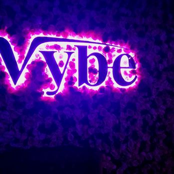 Vybe - The image features a neon sign displaying the word "Vybe" in purple and pink lights, mounted on a textured wall. The sign is illuminated, casting a glow on the surrounding surface, which appears to be decorated with a floral or textured pattern. The lighting and design suggest a lively venue setting, possibly a bar or entertainment space. - Venues & Event Spaces Near Me - Chandler, Arizona