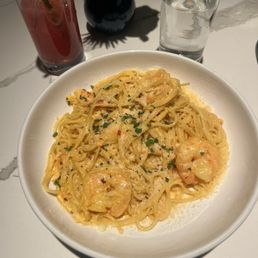 Capellini Marlon - A bowl of shrimp pasta garnished with herbs, served on a white table with drinks and a small vase in the background.