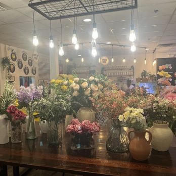 Cake Bar - FLOWERS! - A wooden table holds multiple vases with different types of flowers, set inside a cafe or dessert shop with warm lighting and menu boards in the background. - Food Stands Near Me - Syracuse, New York