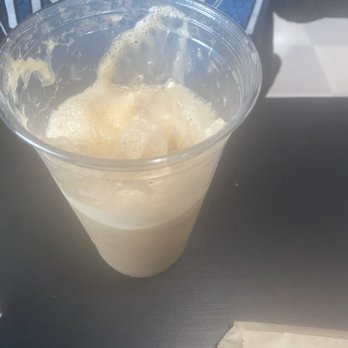 Sweets On Main - Root beer float $5 - A root beer float with vanilla ice cream is served in a clear plastic cup, showing the frothy, creamy beverage inside. The cup is on a dark surface, with part of a napkin visible in the lower right corner. The background includes a person wearing a shirt with a blue and black design. - Ice Cream & Frozen Yogurt Near Me - Fort Wayne, Indiana