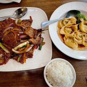 Veggie House - Mongolian Soy Beef; Spicy Dumplings - The image shows a meal with two main dishes and a side of rice on a wooden table. The left plate contains stir-fried vegetables and meat, including onions, green onions, and possibly tofu or pork, with a brown sauce. The right plate has dumplings in a broth, garnished with broccoli. A small bowl of steamed white rice is also present. Silverware is placed on the plates and a napkin is visible at the bottom right. - Vegan Near Me - Chicago, Illinois