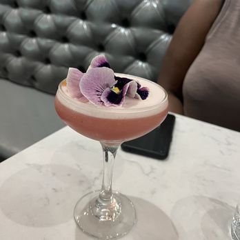 Scarlet Oak - Cherry blossom cocktail - A pink cocktail served in a coupe glass is garnished with purple and pink edible flowers. The drink is on a white marble table, with a smartphone and part of a person wearing a sleeveless top visible in the background. The setting appears to be a restaurant or bar with a tufted, metallic-looking booth behind the table. - New American Near Me - Washington, DC, District of Columbia