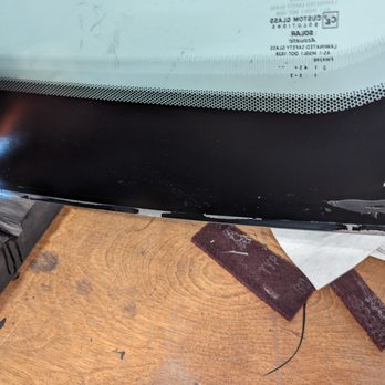 A Better Windshield - The image shows a car windshield with damage along the bottom edge, including chipped glass and cracks. The windshield is placed on a wooden surface, and part of the vehicle's dashboard or interior is visible at the top of the image. The damage appears to be localized to the lower edge, with some peeling or chipping of the glass. - Windshield Installation & Repair Near Me - Monument, Colorado