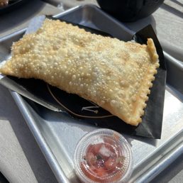 A fried pastry with a bubbly, golden-brown surface is placed on a black paper liner on a metal tray, accompanied by a small container of salsa.
