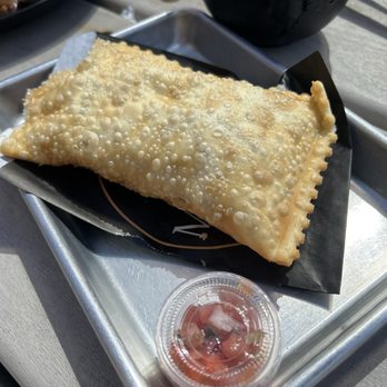 Mama Ana - A fried pastry with a bubbly, golden-brown surface is placed on a black paper liner on a metal tray, accompanied by a small container of salsa. - Breakfast & Brunch Near Me - Somerville, Massachusetts
