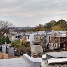 Sealing tight around chimney. - The image shows a residential neighborhood with a row of houses featuring different colors and roof styles, some with solar panels. Trees with sparse foliage are visible, indicating a fall season. In the background, a city skyline with taller buildings can be seen under a cloudy sky. The foreground includes rooftops and a chimney, with some construction materials present.