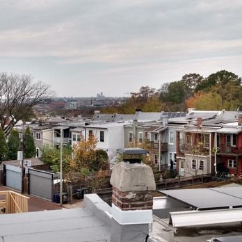 United Roofing - Sealing tight around chimney. - The image shows a residential neighborhood with a row of houses featuring different colors and roof styles, some with solar panels. Trees with sparse foliage are visible, indicating a fall season. In the background, a city skyline with taller buildings can be seen under a cloudy sky. The foreground includes rooftops and a chimney, with some construction materials present. - Roofing Near Me - Washington, DC, District of Columbia