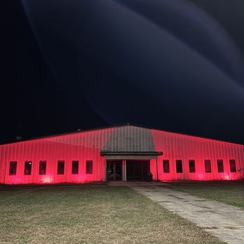 Evil Intentions Haunted House - A metal building with multiple windows is lit with red lights at night, situated on a grassy area with a concrete pathway leading to the entrance. - Haunted Houses Near Me - Maple Park, Illinois