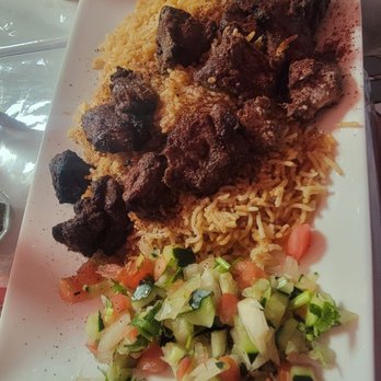 Bamyan Kabob - The image shows a rectangular white plate containing a serving of yellow rice, chunks of grilled meat, and a side of chopped vegetable salad with cucumbers, tomatoes, and onions. The plate is placed on a table, with a glass of beverage visible in the background. The setting suggests a meal at Bamyan Kabob, an Afghan restaurant in Chicago, IL, known for dishes like Kabuli Palaw and Lamb Karahi. - Halal Near Me - Chicago, Illinois