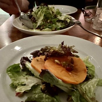 Marina - Persimmon Salad -  salanova lettuce, pomegranate, pistachio, ricotta, pepitas, orange dressing - A white plate contains a salad with sliced persimmon, mixed greens, and nuts, placed on a wooden table. In the background, another salad with greens and a glass of water are visible, indicating a dining setting. - Italian Near Me - Pasadena, California