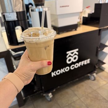 Koko Coffee - A person holds a plastic cup filled with iced coffee, with a straw, in front of a Koko Coffee counter. The counter has a black front with the Koko Coffee logo, and coffee-making equipment, including a grinder and a coffee machine, are visible in the background. The setting appears to be inside a coffee shop located on the second floor of a mall. - Coffee & Tea Near Me - Cambridge, Massachusetts
