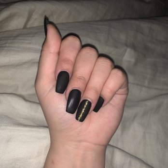 Orchid Nails - Matte Black Acrylic Set - A close-up of a hand with five fingers, showcasing matte black acrylic nails. The ring finger has a gold stripe detail running vertically down the center. The background consists of a neutral-colored fabric, possibly a bed or blanket. The nails are well-manicured and the image emphasizes the nail design and finish. - Nail Salons Near Me - Lisle, Illinois