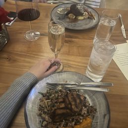 Mushroom risotto (wild rice because changes to supply) - A wooden dining table features two plates of food, one with grilled meat served over rice and vegetables, and the other with a dark meat dish accompanied by mashed potatoes. There are three glasses: one with red wine, one with champagne, and one with water and ice. A person wearing a gray sweater is holding a champagne flute, and another person in a striped shirt is seated across, with a plate of food in front of them. A menu is partially visible on the right side of the table.