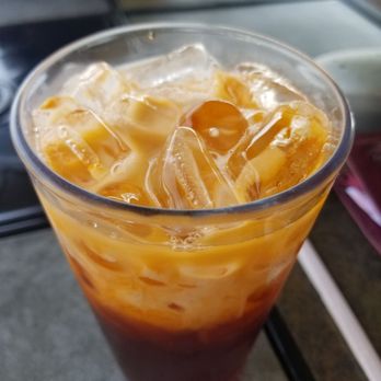 Thai Lada - Iced Thai Milk Tea - A clear, tall glass contains iced Thai milk tea with visible layers of dark tea and creamy milk, topped with ice cubes. The beverage appears cold and refreshing, with a mix of orange and brown hues, placed on a dark surface with a blurred background. - Thai Near Me - Austin, Texas