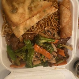 Egg Roll - A foam takeout container holds beef with mixed vegetables, an egg roll, fried wontons, and stir-fried noodles, typical of Chinese cuisine.