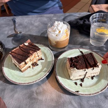 Saluti’s - Two plates with layered chocolate and vanilla cakes drizzled with chocolate, a dessert with whipped cream, and a glass of water are on a table at a restaurant. - Italian Near Me - Jackson, California