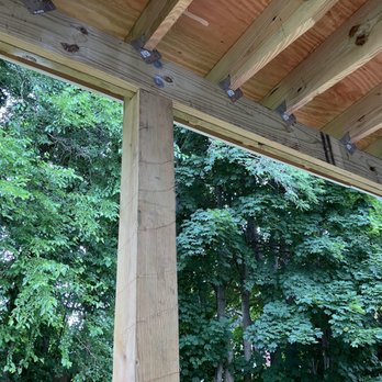 DTails Paint with Class - Deck roof - The image shows a partially built wooden structure with vertical and horizontal beams, with green trees in the background. - Painters Near Me