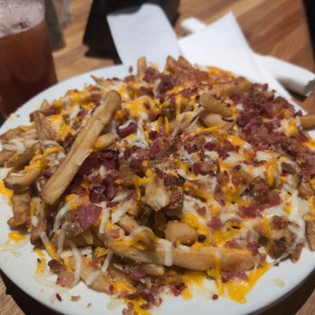 Cheddar's Scratch Kitchen - Cheese fries appetizer - A large white plate holds loaded fries topped with melted cheese, crispy bacon bits, and cooked chicken pieces. The plate is placed on a wooden table, with a glass of iced beverage, a napkin, and utensils visible in the background. The setting appears to be a casual dining restaurant. - American Near Me - York, Pennsylvania