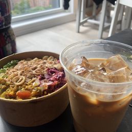 Vegetarian congee and Vietnamese Iced Coffee - A bowl of vegetarian congee containing mixed vegetables and toppings is positioned on a table near a large window, with a view of city buildings in the background. Next to the congee, there is a clear plastic cup filled with iced Vietnamese coffee, featuring ice cubes and a creamy, coffee-colored liquid. The setting appears to be indoors, possibly in a cafe, with chairs and a table visible in the background.