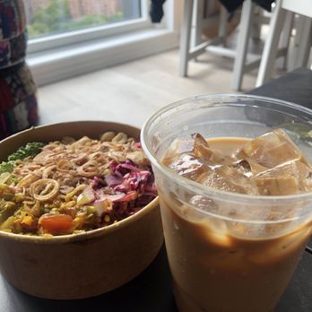 Maya Congee Cafe - Vegetarian congee and Vietnamese Iced Coffee - A bowl of vegetarian congee containing mixed vegetables and toppings is positioned on a table near a large window, with a view of city buildings in the background. Next to the congee, there is a clear plastic cup filled with iced Vietnamese coffee, featuring ice cubes and a creamy, coffee-colored liquid. The setting appears to be indoors, possibly in a cafe, with chairs and a table visible in the background. - Coffee & Tea Near Me - Brooklyn, New York