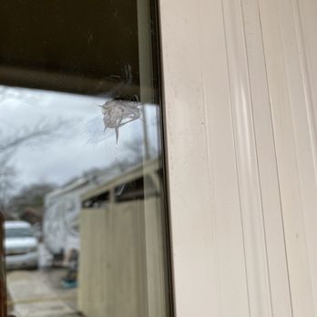 AtticDr - a window with a hole in it - The image shows a window with a small, damaged area and a crack near the center. The window reflects an outdoor scene with vehicles and a cloudy sky. The window frame is white, and the surrounding wall has vertical siding. The damage appears to be a chip or hole in the glass, with a crack extending from it. - General Contractors Near Me - Austin, Texas