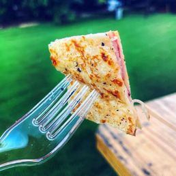 Ham and Cheese Crepe - A slice of ham and cheese crepe is pierced on a clear plastic fork, held outdoors near a wooden surface. The crepe has a golden-brown exterior with visible black pepper and seasoning, and pink ham and melted cheese are visible inside. The background features a grassy area with trees and a blurred outdoor setting, indicating a casual outdoor dining environment.