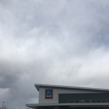 ALDI - A parking lot with multiple cars parked in front of an ALDI supermarket. The store's sign is visible on the building's facade, which has a modern architectural design with large windows. The sky above is overcast with gray clouds, indicating cloudy weather. - Grocery Near Me - South Windsor, Connecticut