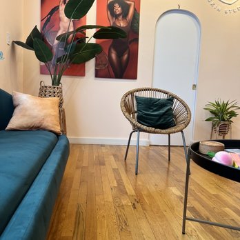 PeachFuzz Laser Studio - A skincare studio interior features artwork of women, potted plants, a wicker chair with a cushion, and a teal sofa with a pillow. - Laser Hair Removal Near Me - Brooklyn, New York