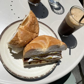 Caffe Social - A plate holds a sandwich with layered ingredients including eggs and possibly meat, served with a piece of bread. On the white outdoor table, there are two glasses of iced coffee or similar beverages, one with foam and a straw, and another dark drink. A glass of water is also present, with sunlight casting shadows on the table. - American Near Me - Norwalk, Connecticut