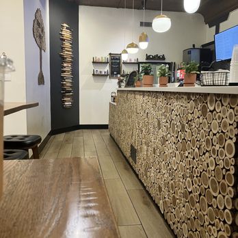 Urban Pine Eatery - Service counter - The image shows the interior of a cafe or eatery with a service counter covered in a wood log pattern. Above the counter, there are five hanging pendant lights with white and gold accents. Behind the counter, there are shelves with small potted plants, nail polish bottles, and other items. The wall to the left has a decorative piece resembling a tree made of wood or similar material, and a vertical display of wooden sticks or logs. The floor is tiled, and part of a wooden table and a stool are visible in the foreground. The ceiling is dark with track lighting and an air conditioning unit. - Pizza Near Me - Flagstaff, Arizona
