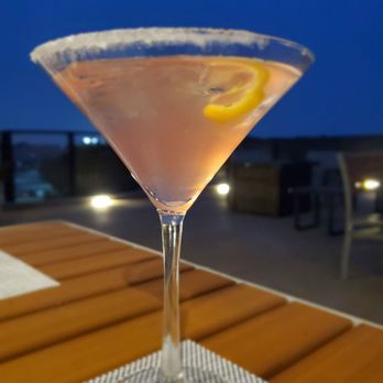 Devil's Kitchen - A clear martini glass contains a pinkish cocktail garnished with a lemon slice. The glass is placed on a wooden table with a woven placemat. The background shows an outdoor setting with chairs, tables, and a darkening sky, indicating evening or night. - New American Near Me - Grand Junction, Colorado
