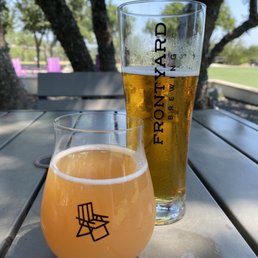 Raspberry Lemonade - Two glasses of beer are positioned on an outdoor metal table at Frontyard Brewing in Spicewood, TX. The taller glass contains a golden beer with condensation, while the shorter glass holds a cloudy, pale beer. The background features trees, a park, and a clear blue sky, indicating a sunny day. The branding on the glasses includes 'FRONTYARD BREWING' on the tall glass and a stylized chair logo on the shorter glass. The setting appears to be a spacious outdoor area with shaded seating and natural surroundings.