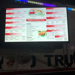 The image features a digital menu board for El Compa Mexican food truck, illuminated with colorful LED lights at the top. The menu lists various traditional Mexican dishes such as tacos, burritos, gorditas, quesadillas, and sides, with prices ranging from $3.50 to $15.50. The menu also includes options for regular and premium meat, and a kid's menu. The background is dark, emphasizing the bright display of the menu.