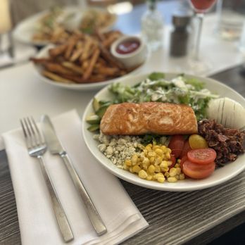 501 Local - A white plate contains a serving of cooked salmon, a mixed salad with lettuce and dressing, yellow corn kernels, halved cherry tomatoes, and a boiled egg. The plate is placed on a dark wood table with a white napkin, fork, and knife. In the background, there are French fries with ketchup, a drink in a martini glass, and a blurred setting suggesting a restaurant environment. - New American Near Me - Winnetka, Illinois